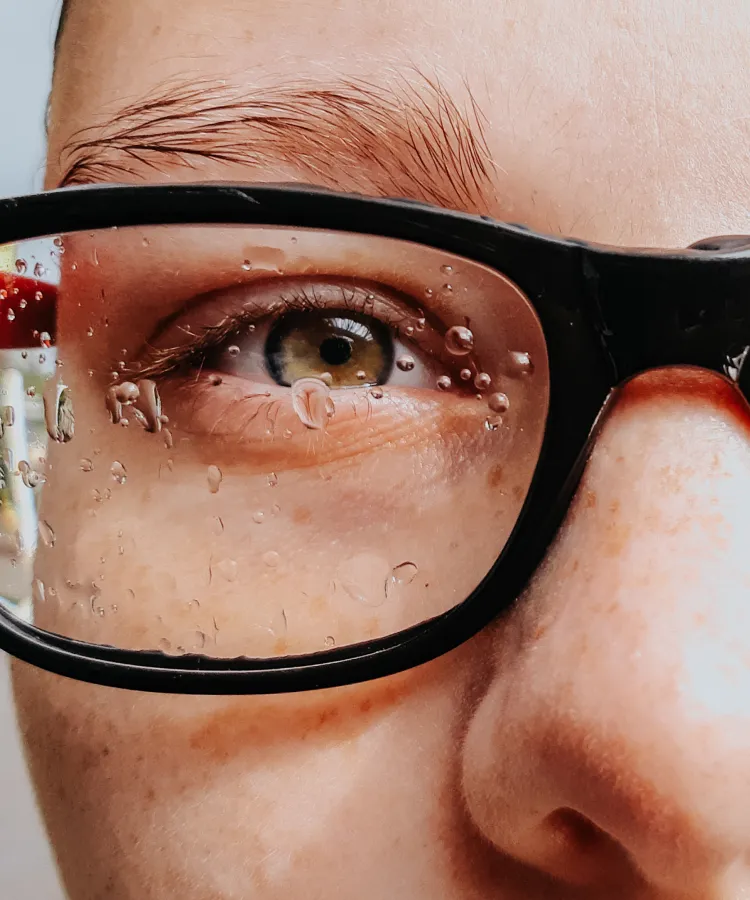 Close-up of a person’s face showing their eye behind black-framed glasses with water droplets on the lenses.