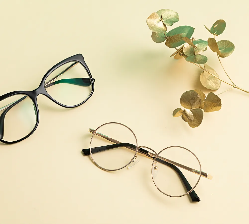 Two pairs of eyeglasses, one black rectangular and one round metal frame, placed on a pale yellow background next to a branch with golden and green leaves.