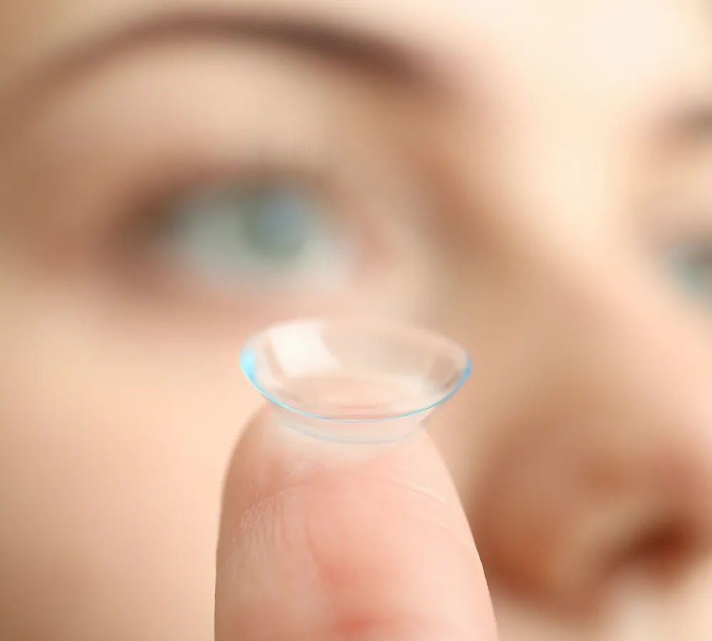 Close-up of a contact lens balanced on the tip of a finger with a blurred face in the background.