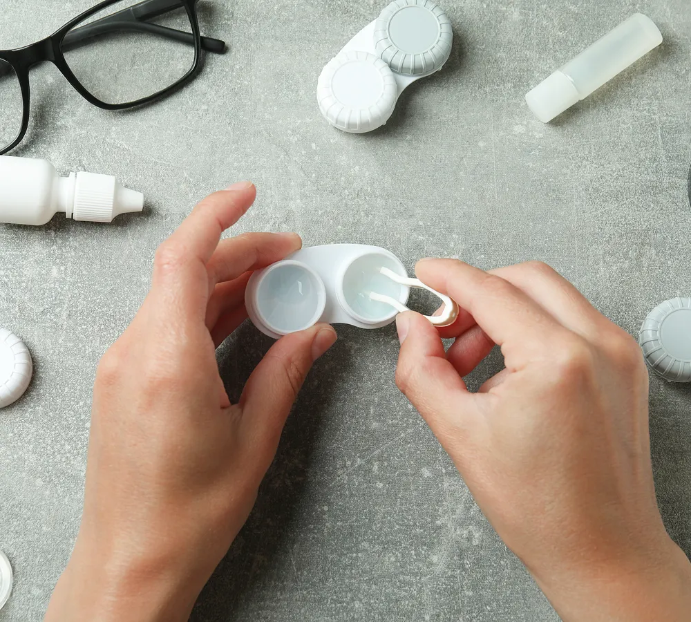 Hands using tweezers to place a contact lens into a white contact lens case on a gray surface with eyeglasses and eye care bottles nearby.