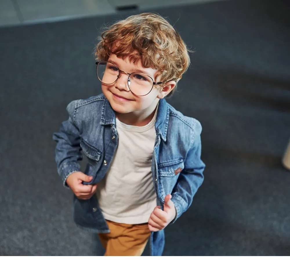 Smiling young boy with curly hair wearing large glasses, a denim jacket, and mustard pants.