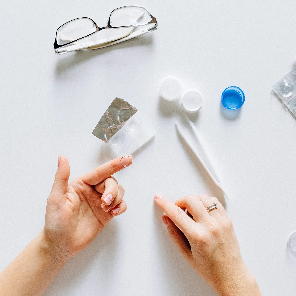 Person holding a contact lens on the fingertip with contact lens case, tweezers, and eyeglasses on a white surface.