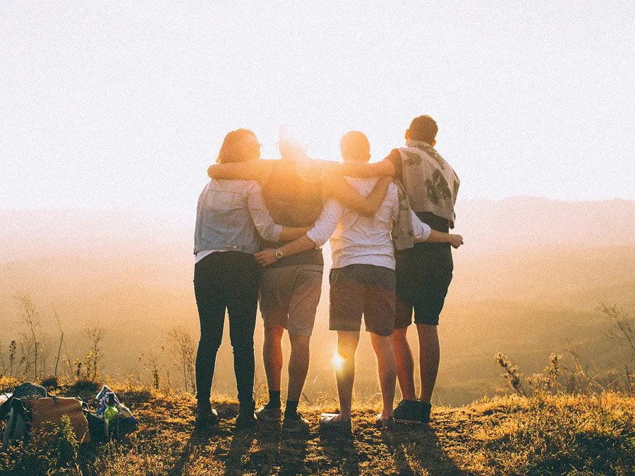 Four friends standing arm in arm on a hilltop facing a golden sunset with backpacks on the ground.