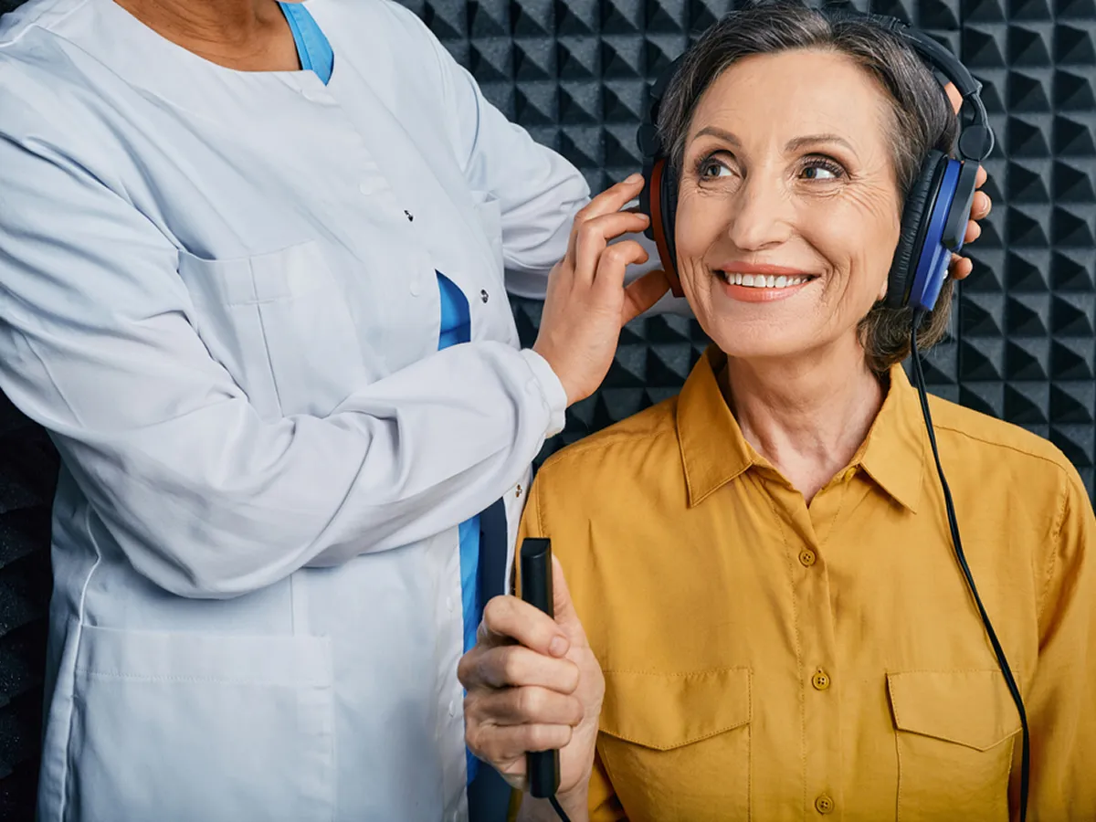 An elderly woman smiling while wearing headphones as a healthcare professional adjusts them in a soundproof room.