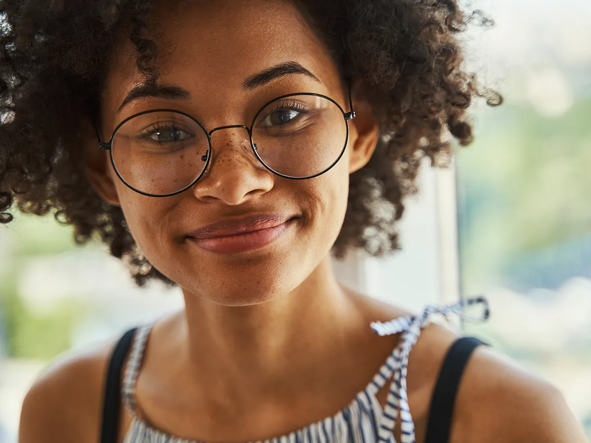 Close-up of a smiling young woman with curly hair and round glasses wearing a striped top with a bow on the shoulder.