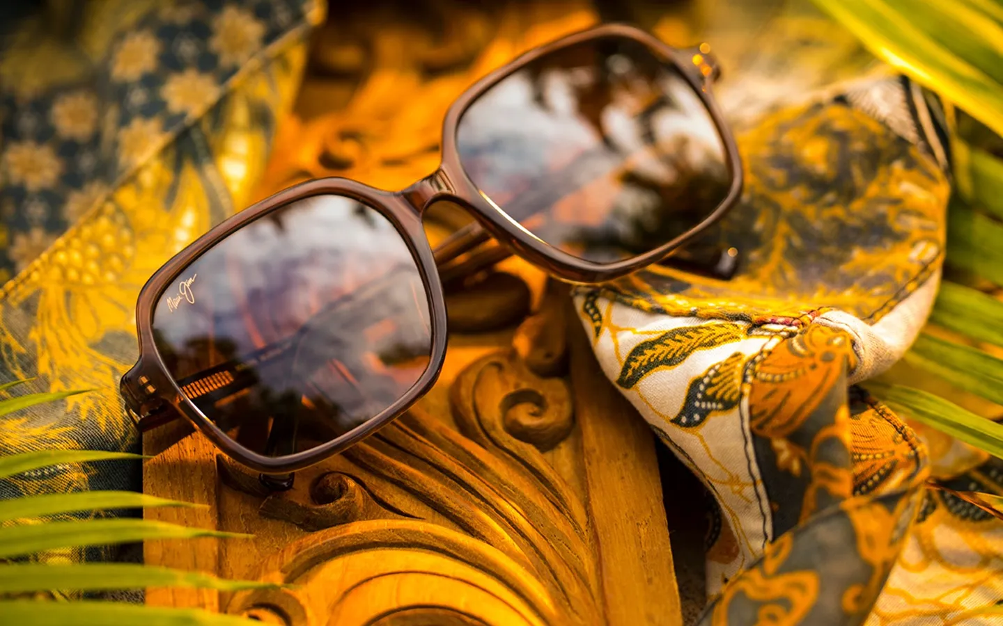 Maui Jim sunglasses with dark lenses resting on carved wooden surface next to a yellow and blue patterned cloth.