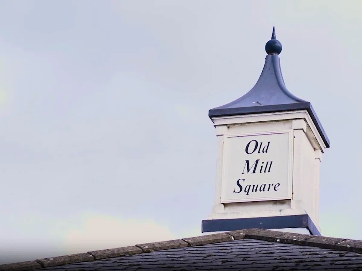 White cupola with dark blue pointed roof and a sign reading 'Old Mill Square' on top of a building roof.