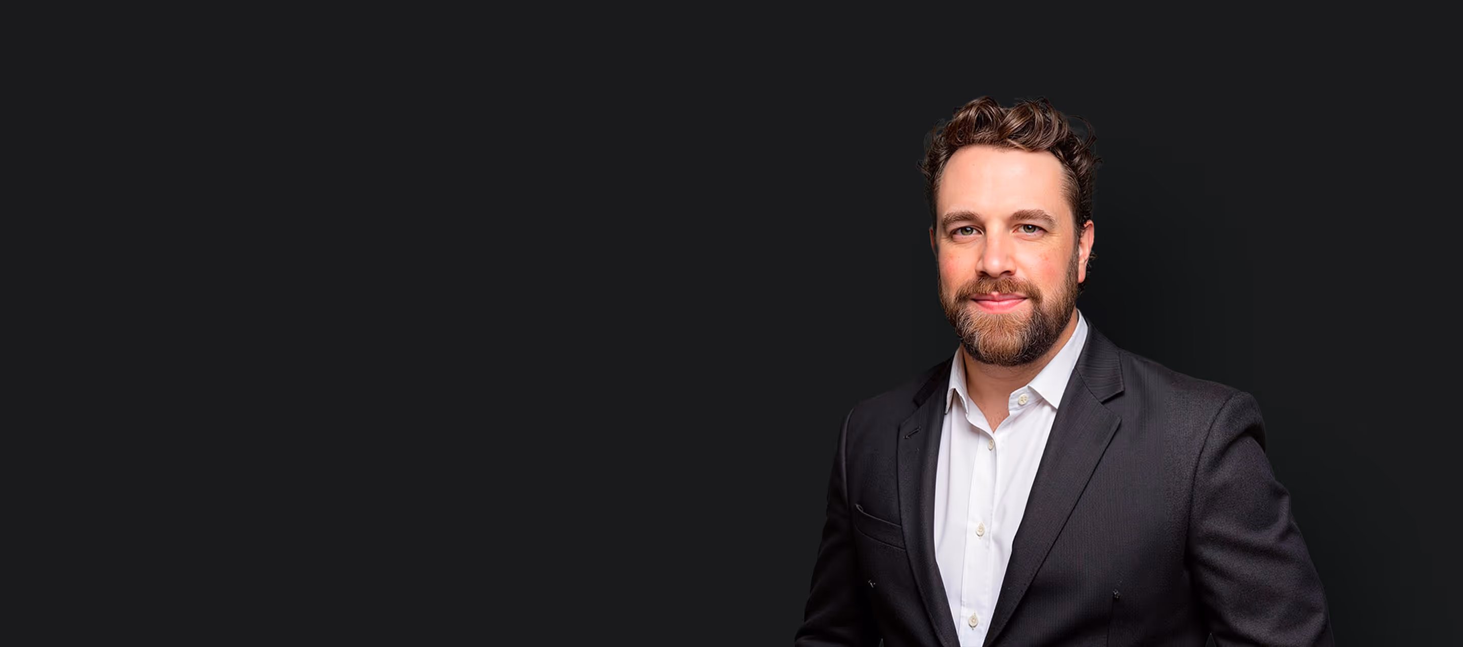 Smiling man with beard wearing a black suit jacket and white shirt against dark background.
