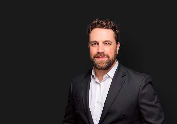 Portrait of a smiling man with beard and curly hair wearing a black suit jacket and white shirt against a dark background.