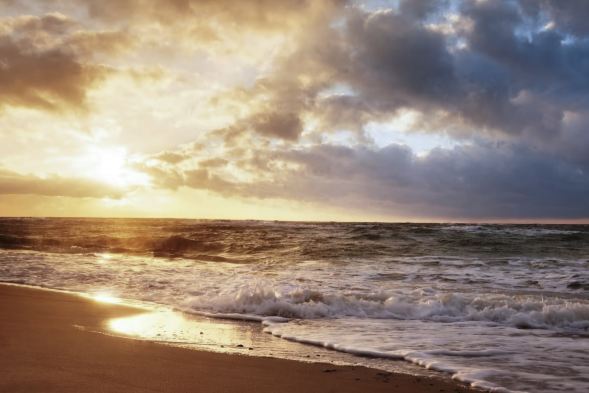 Calm ocean waves washing onto a black sand beach with a mountain range reflected in the water at sunset.