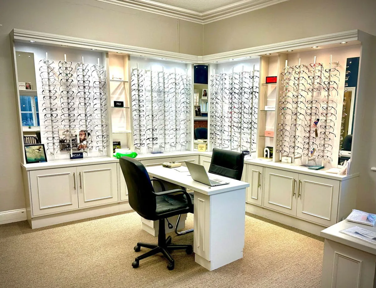 Bright optician's office with a white desk, two black chairs, and wall displays filled with diverse eyeglass frames.