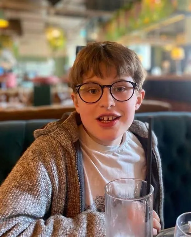 Young boy with glasses and brown hair sitting at a table, holding a large glass in a cozy indoor setting.