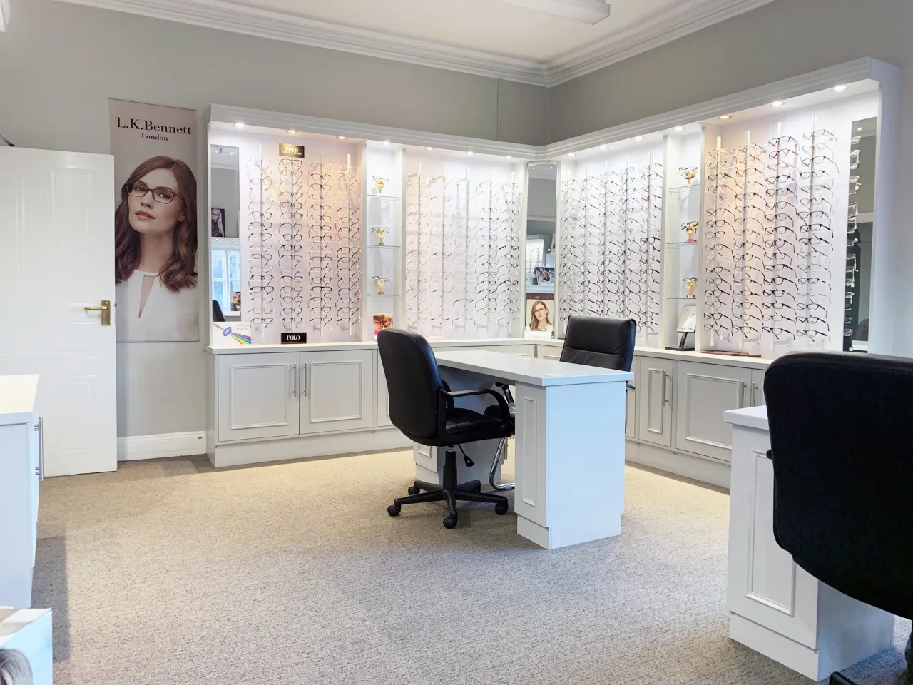 Interior of an optical store with white display cabinets filled with numerous eyeglasses and two black office chairs around a white desk.