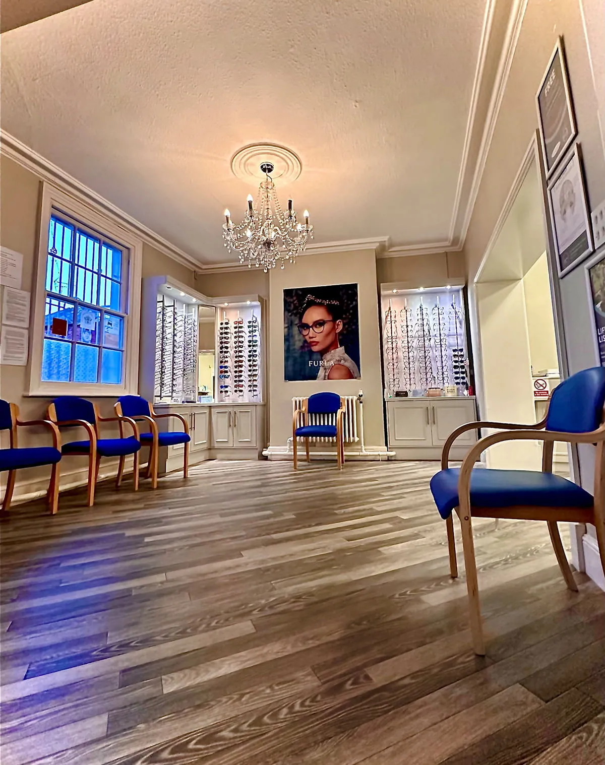 Optician waiting room with wooden floor, blue cushioned chairs, eyeglass displays, and a chandelier.