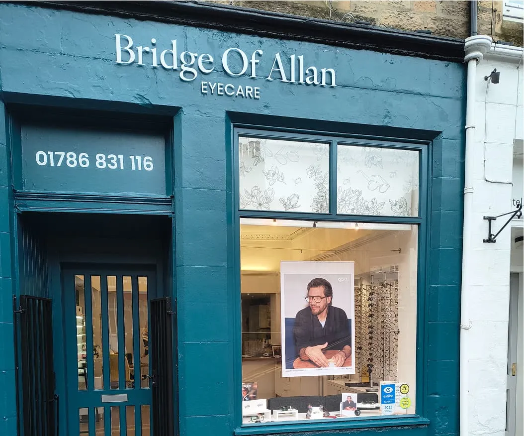 Front view of Bridge Of Allan Eyecare shop with a teal painted exterior, phone number 01786 831 116, and a window display featuring a poster of a man wearing glasses.