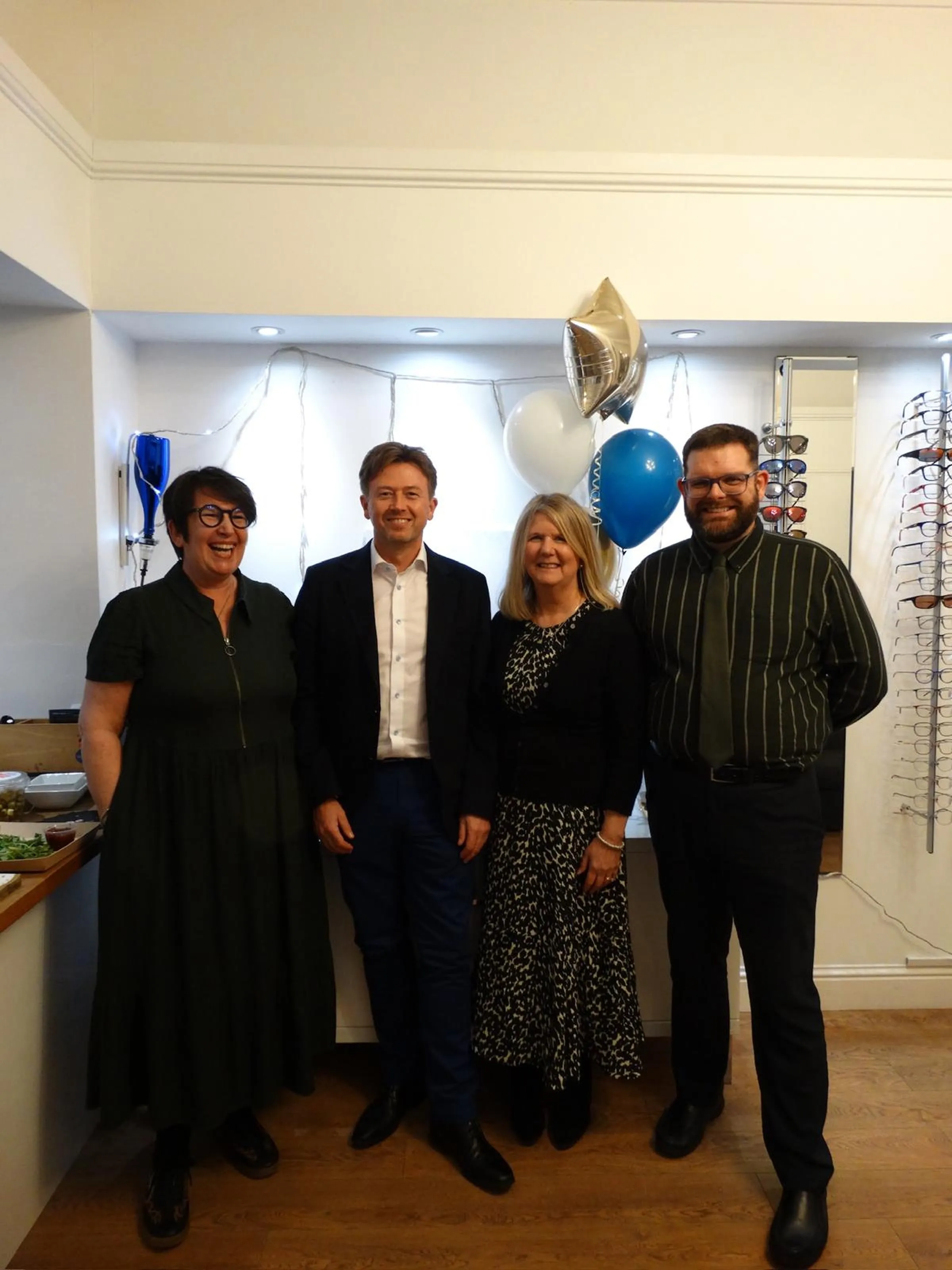 Four adults standing side by side indoors with blue, white, and silver balloons in the background and a display of eyeglasses on the wall.