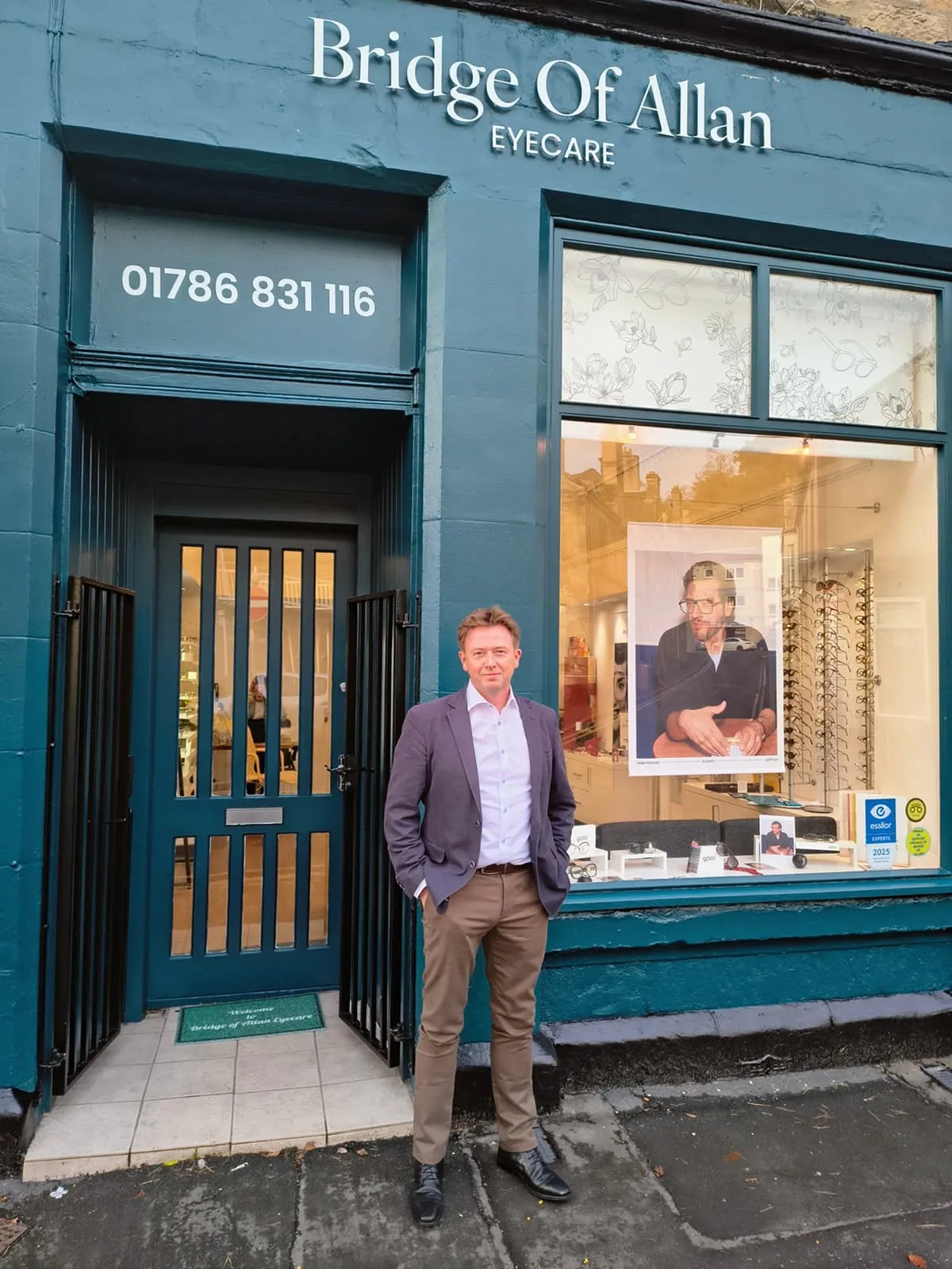 Man in a blazer and khaki pants standing in front of Bridge Of Allan Eyecare shop with eyeglasses displayed in the window.