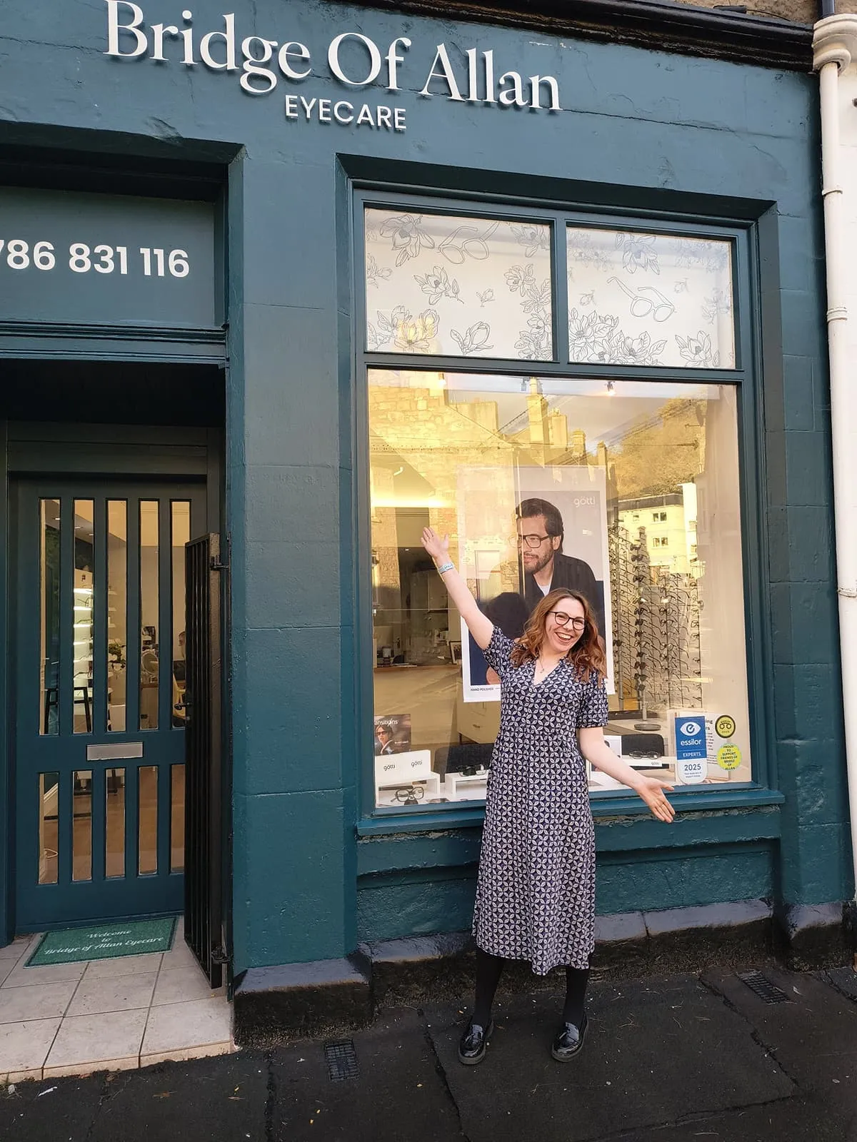 Smiling woman with glasses stands outside Bridge Of Allan Eyecare shop, gesturing to the window display.