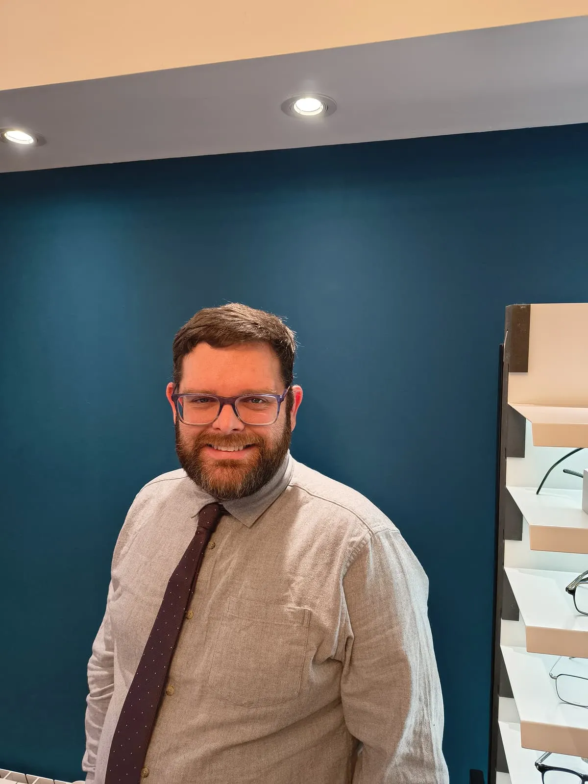 Smiling man with glasses, beard, wearing a light gray shirt and dark tie, standing in front of a blue wall with glasses displayed on shelves to the right.