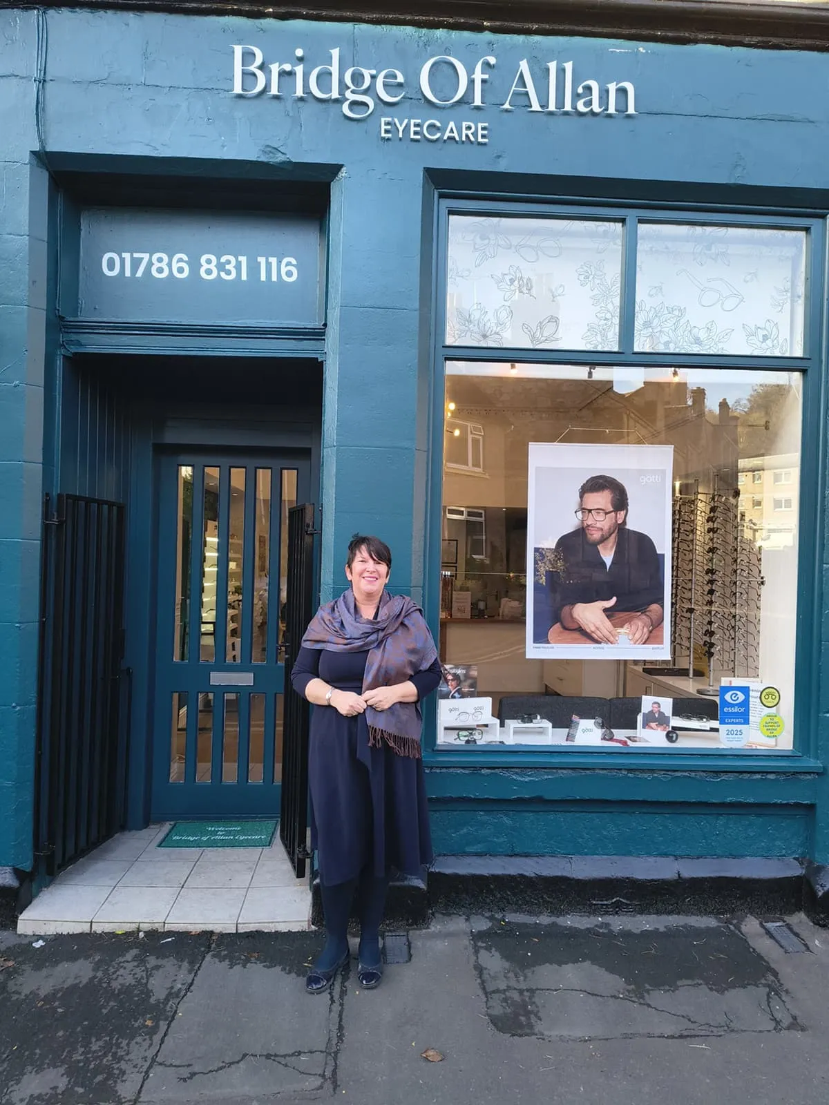 Woman standing smiling in front of Bridge Of Allan Eyecare shop with a poster of a man wearing glasses in the window.