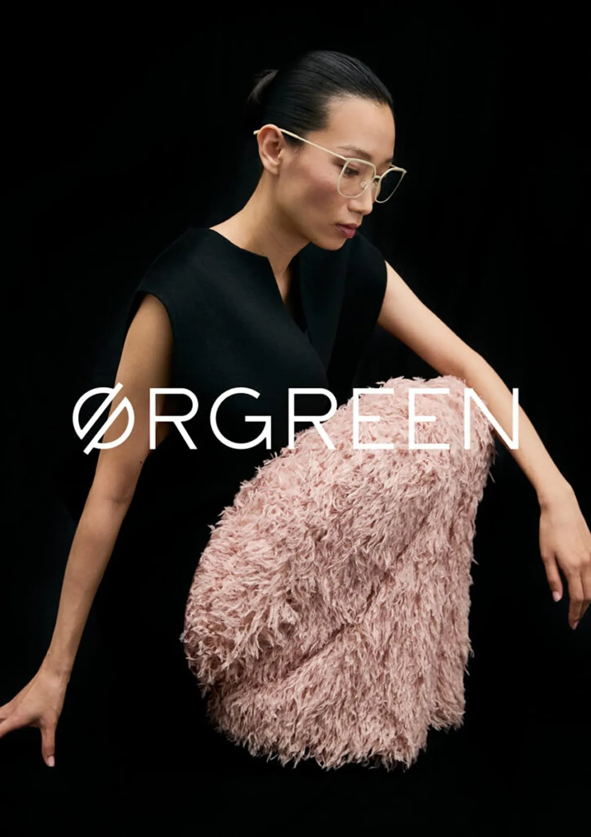 Person with short black hair and gold frame glasses wearing a sleeveless black top and a textured pink skirt against a black background with the ØRGREEN logo overlay.