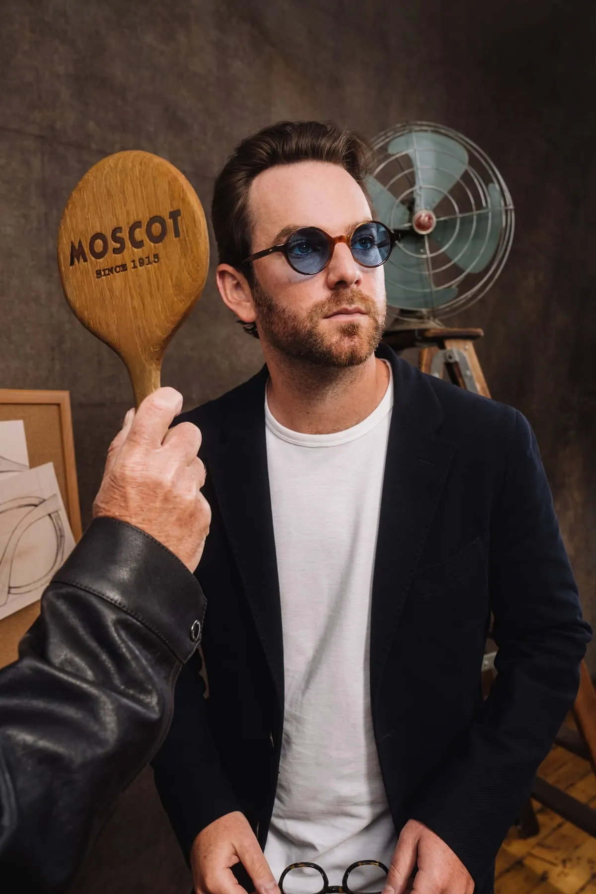 Man wearing round tortoiseshell sunglasses with blue lenses poses as a wooden paddle with MOSCOT logo is held beside his face.