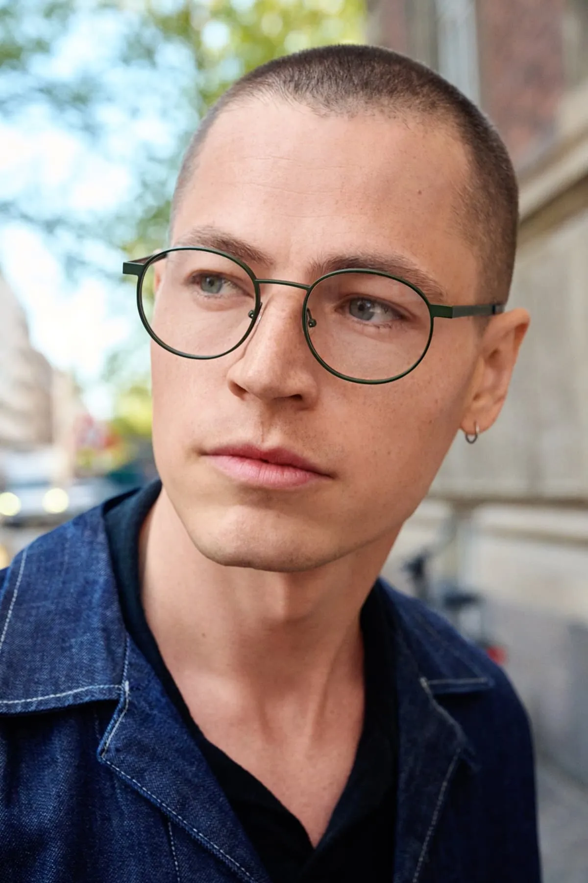 Close-up of a young man with short hair wearing round glasses and a denim jacket, looking to the side outdoors.