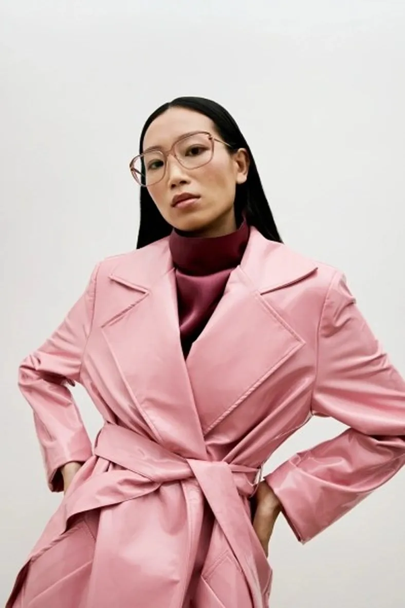 Woman with long black hair wearing large pink transparent glasses, a maroon turtleneck, and a pink belted trench coat.
