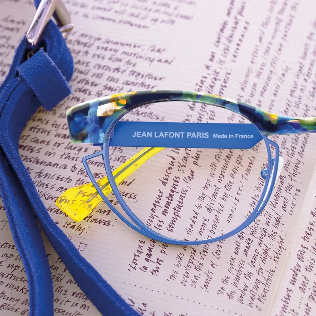 Colorful Jean Lafont Paris eyeglass frame with yellow temple tips resting on handwritten text, next to a blue suede strap.