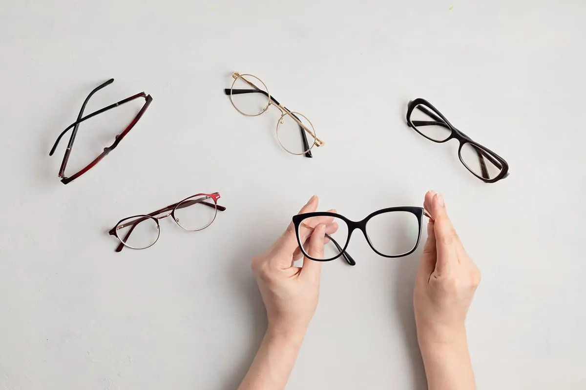Two hands holding a pair of black cat-eye eyeglasses with four other pairs of eyeglasses laid out on a white surface.