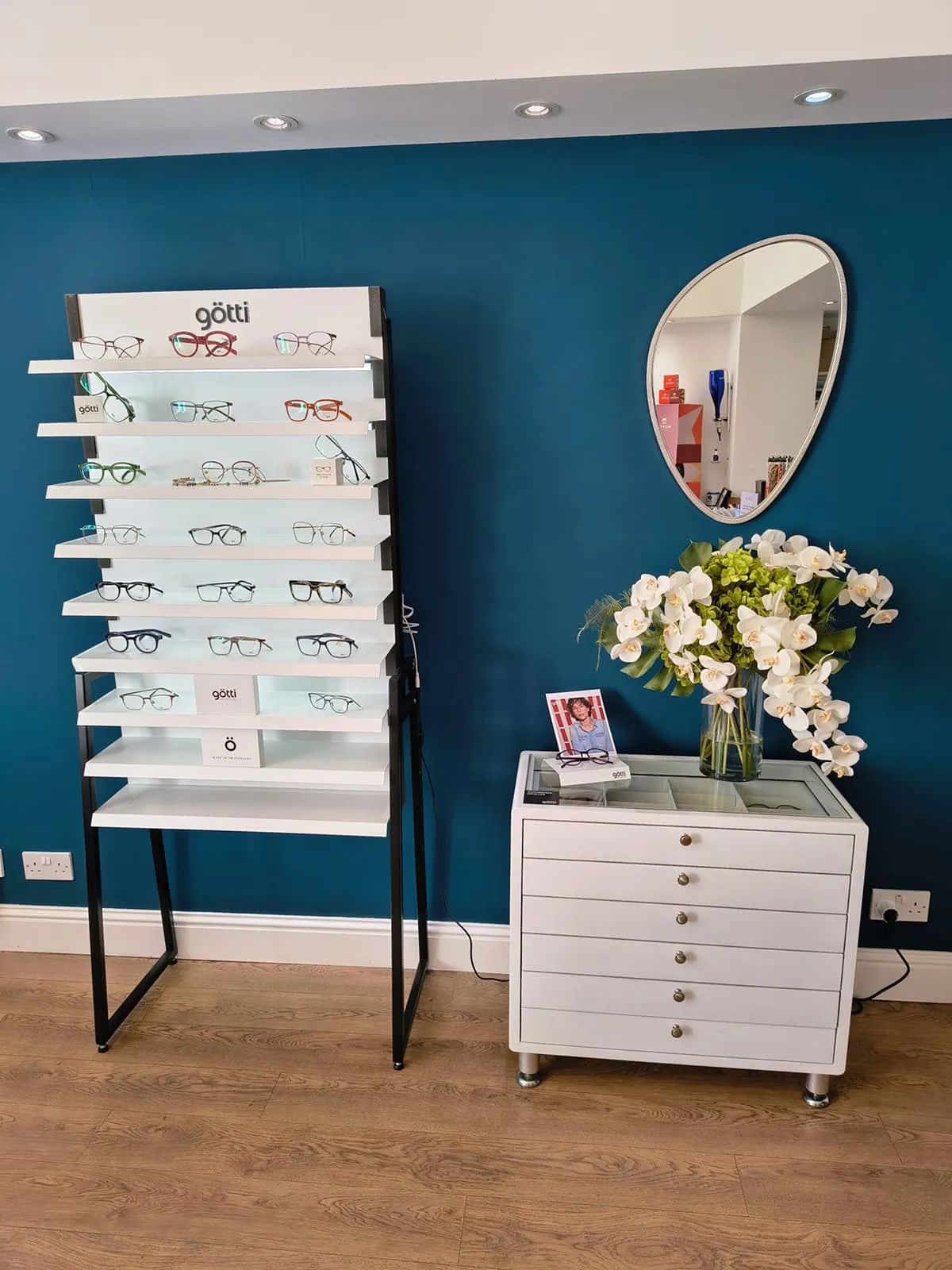 Display stand with multiple pairs of eyeglasses next to a white cabinet with drawers, a vase of white and green flowers, and an oval mirror on a teal wall.