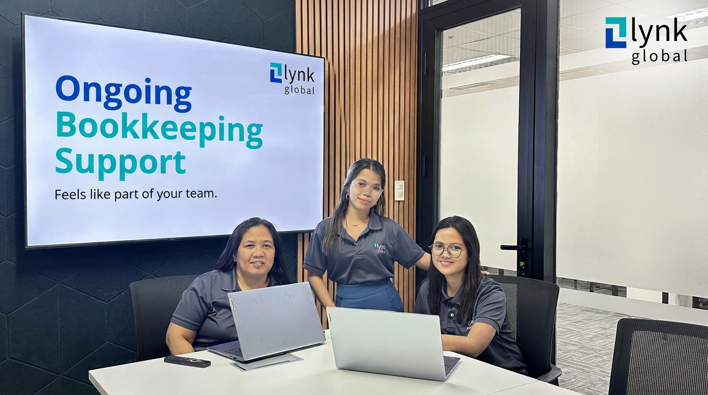 Three women in matching gray polo shirts sitting and standing at a conference table with laptops, next to a screen displaying 'Ongoing Bookkeeping Support Feels like part of your team.' and the Lynk Global logo.
