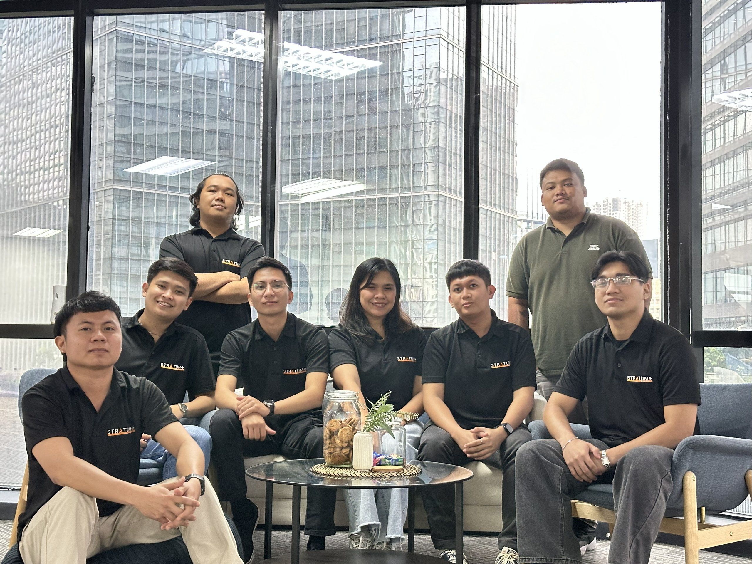 Group of eight people, mostly wearing black polo shirts with STRATUM logos, sitting and standing in a modern office with large windows showing city buildings.