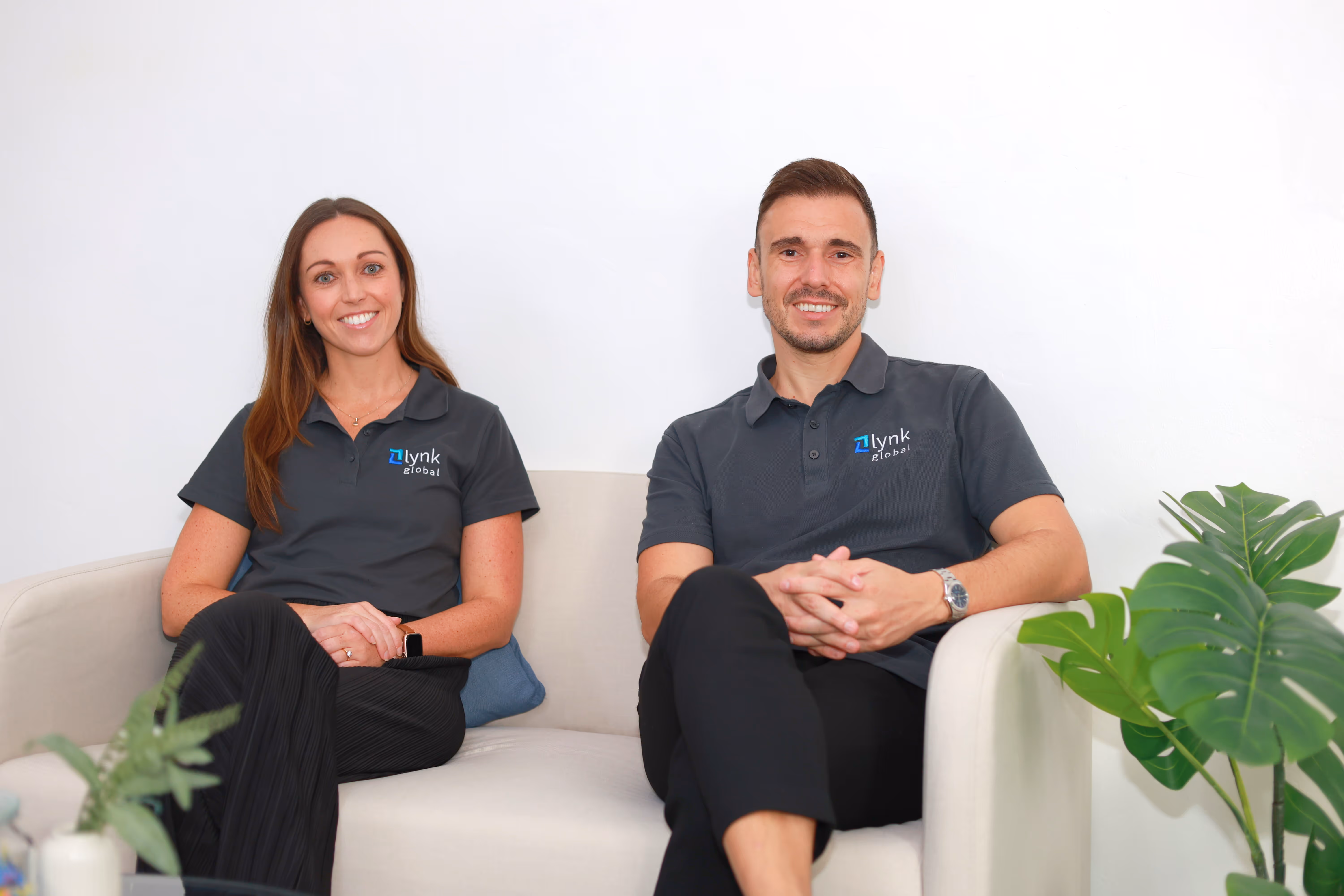 Man and woman wearing Lynk Global shirts sitting on a white couch smiling, with a green plant on the right.