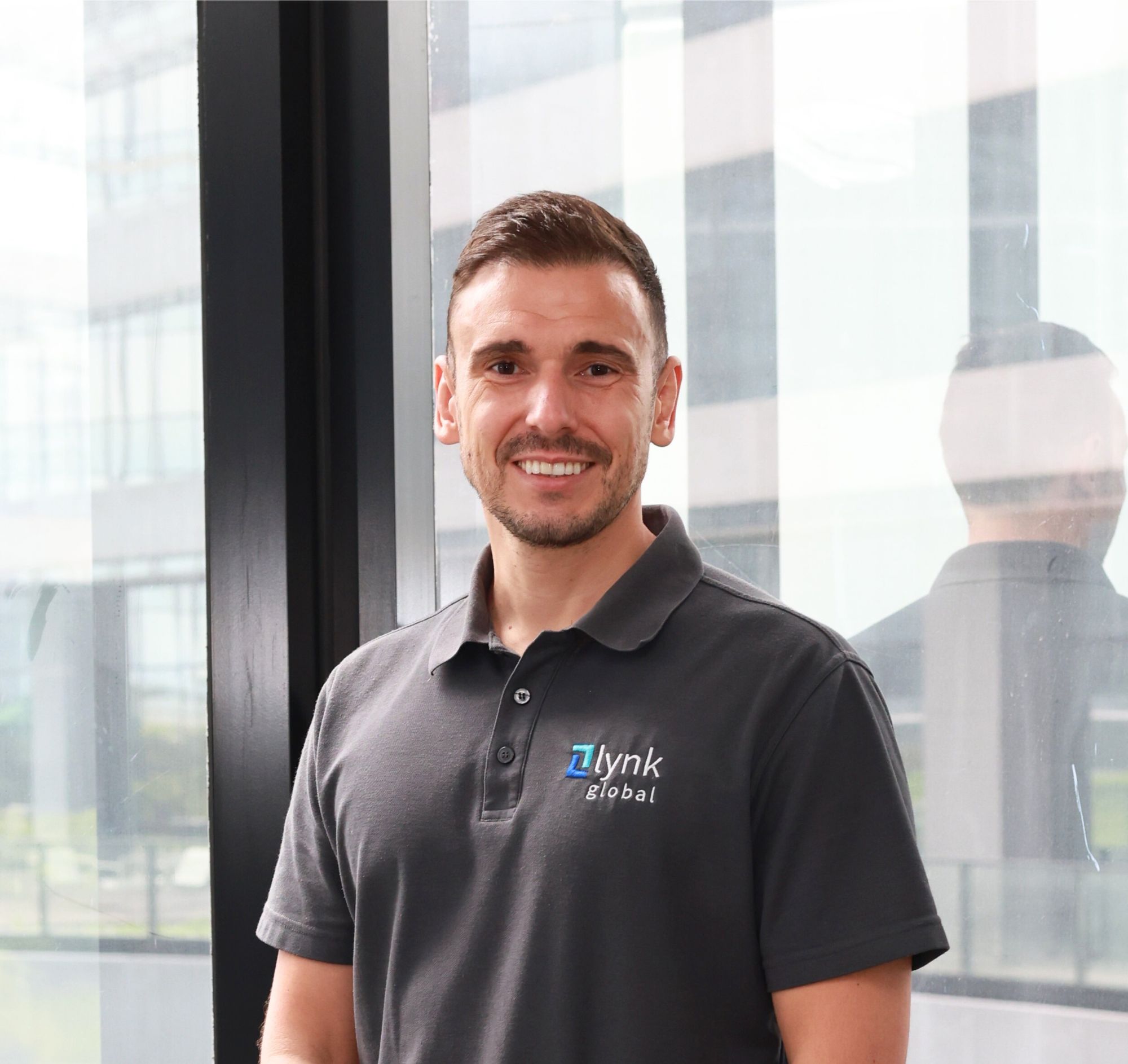 Smiling man wearing a dark polo shirt with 'Lynk Global' logo standing indoors near a glass wall.