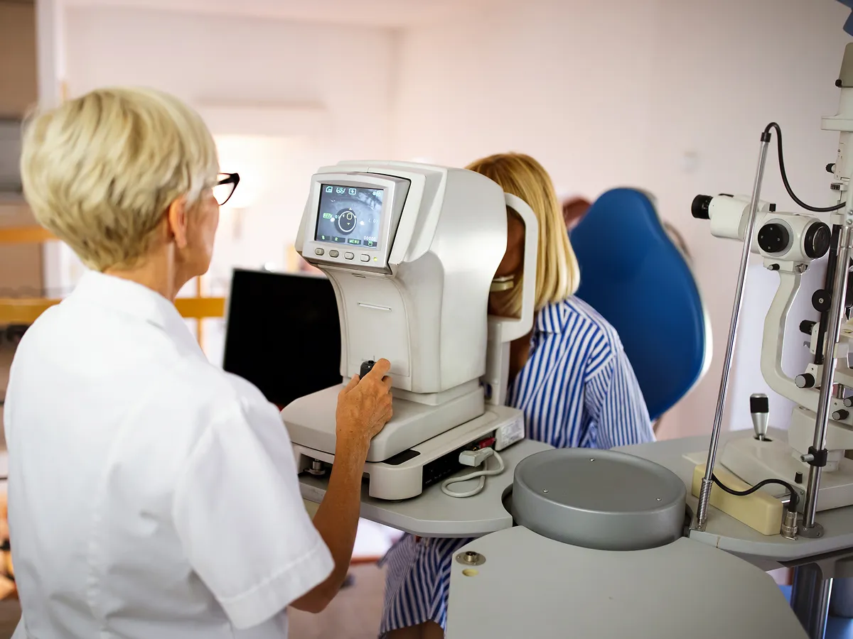 Optometrist adjusting a machine to perform an eye test on a woman wearing a striped shirt.