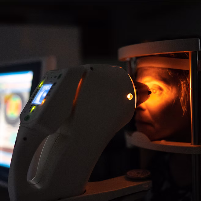 Person undergoing an eye exam with a retinal scanner emitting orange light in a dark room.