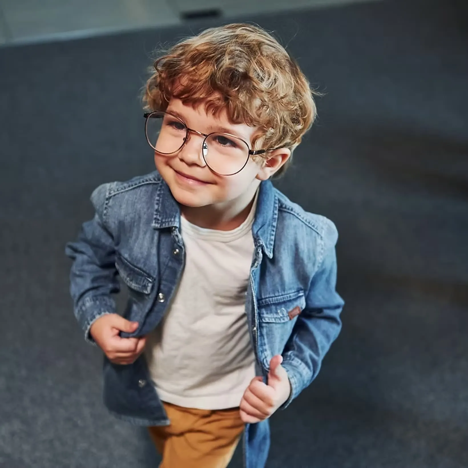 Smiling young boy with curly hair wearing large glasses, a denim jacket, and mustard pants.