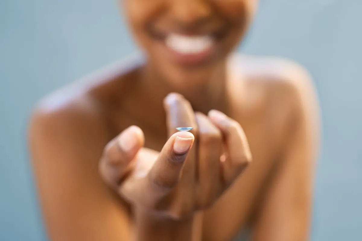 Close-up of a contact lens balanced on the tip of a finger with a blurred face in the background.