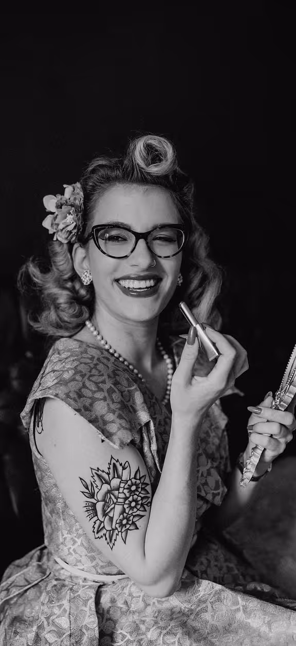 Schwarz-weiß-Porträt einer lächelnden Frau mit Retro-Frisur, Brille, Perlenkette und blumenförmigem Tattoo am Oberarm, die Lippenstift hält und sich in einem Handspiegel betrachtet.