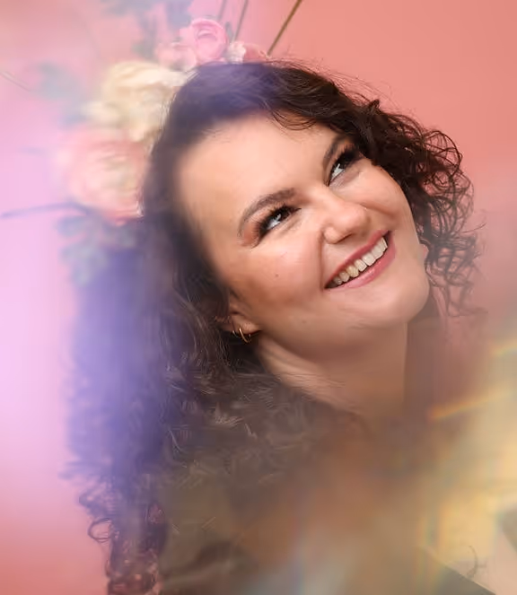 Frau mit lockigem Haar lächelt und schaut nach oben, vor einem rosa Hintergrund mit verschwommenen Licht- und Blütenreflexen.
