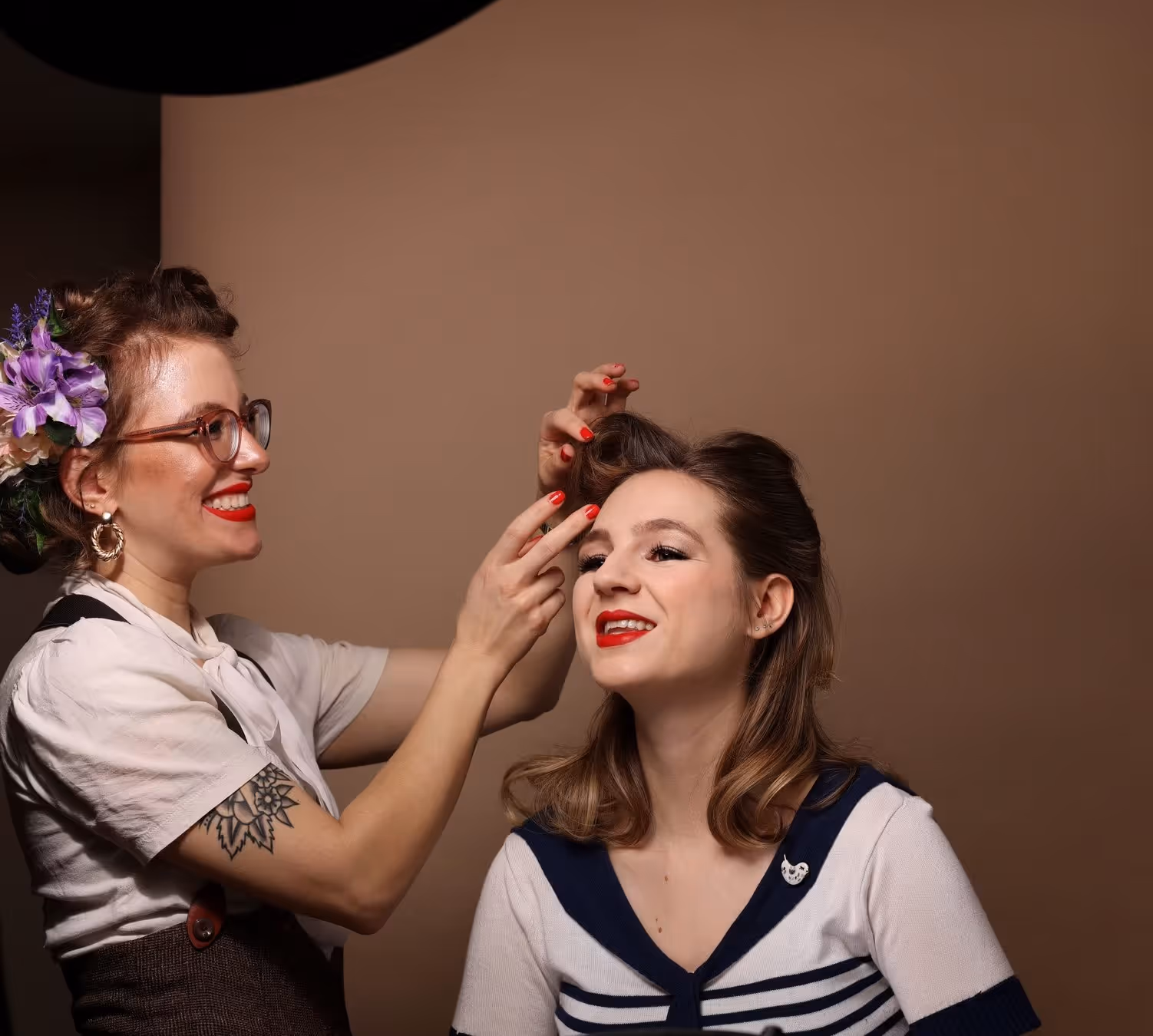 Friseurin mit Blumen im Haar stylt lachend das Haar einer Frau mit rot geschminkten Lippen vor braunem Hintergrund.