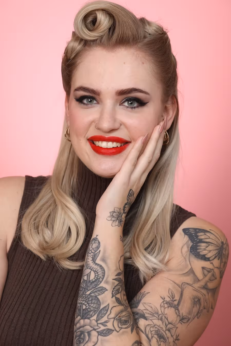 Porträt einer lächelnden blonden Frau mit rotem Lippenstift, vintage Frisur, schwarzen Augen-Make-up und tätowierten Armen vor rosa Hintergrund.