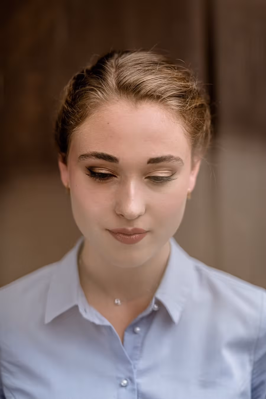 Porträt einer jungen Frau mit hochgesteckten blonden Haaren, dezentem Make-up und hellblauer Bluse, die nach unten blickt.