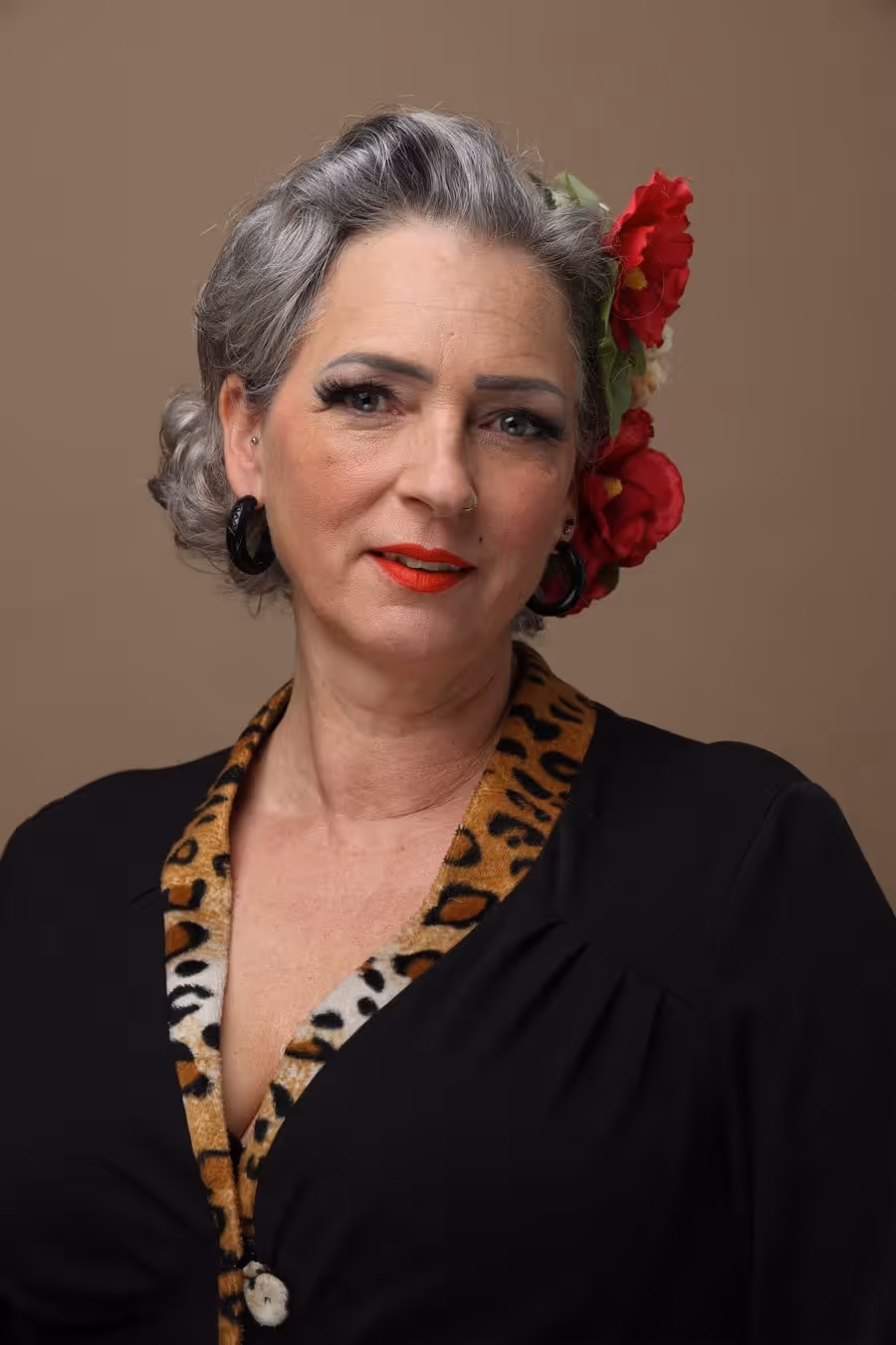 Ältere Frau mit grauem Haar, roten Blumen im Haar, rotem Lippenstift und schwarzem Kleid mit Leopardenmuster-Kragen.