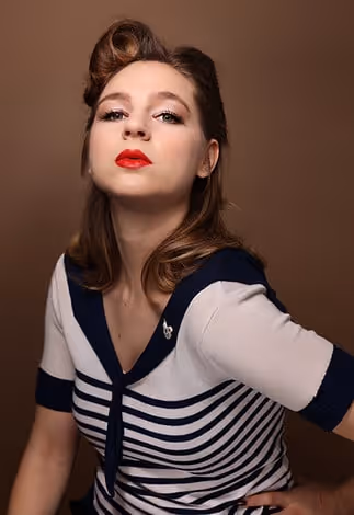 Porträt einer Frau mit Retro-Frisur und rotem Lippenstift, die ein weißes und blau gestreiftes Oberteil trägt, vor braunem Hintergrund.
