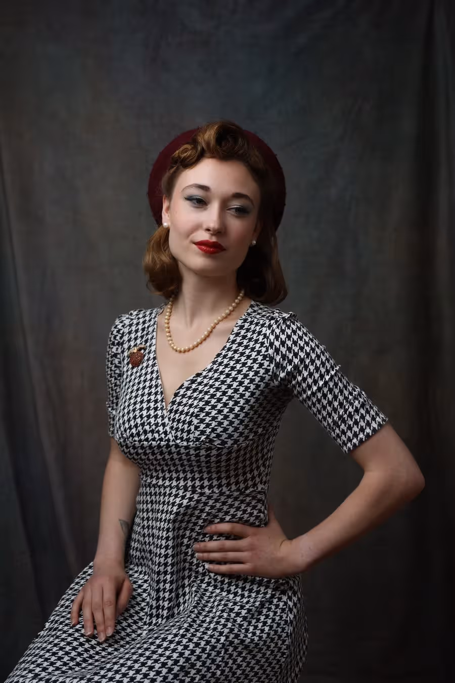 Frau mit Retro-Frisur trägt ein schwarz-weißes Hahnentritt-Kleid, Perlenkette und roten Hut vor dunklem Hintergrund.