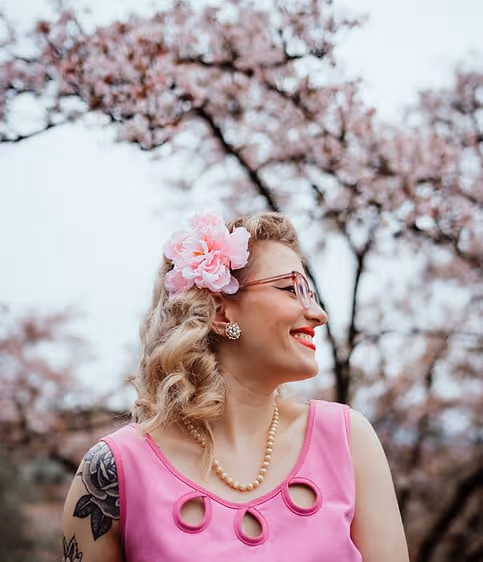 Frau mit rosa Blume im Haar, rosa Kleid und Perlenkette lächelt vor blühenden Kirschblütenbäumen.