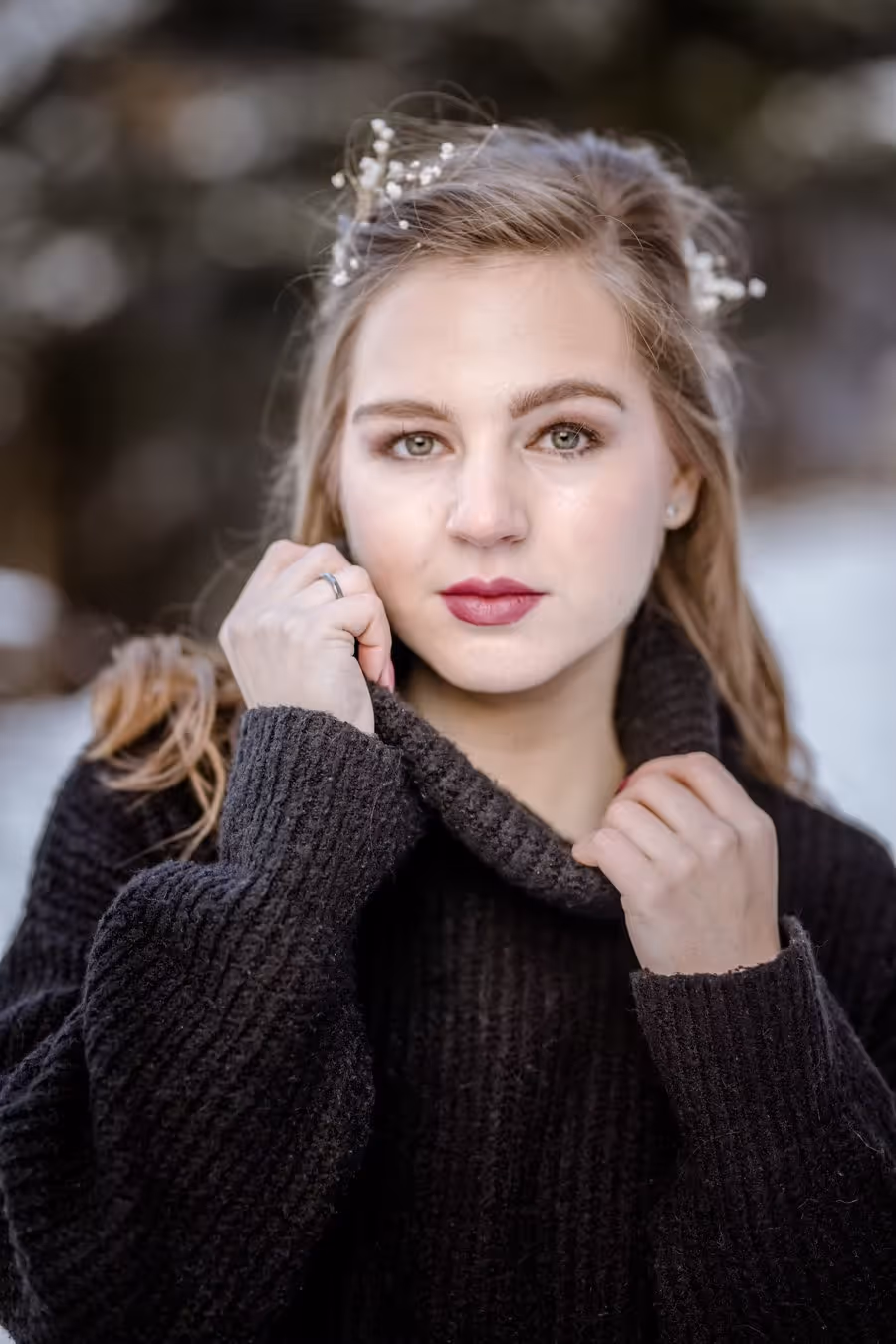 Junge Frau mit blonden Haaren, roten Lippen und Blumen im Haar, trägt einen schwarzen Strickpullover mit hohem Kragen.