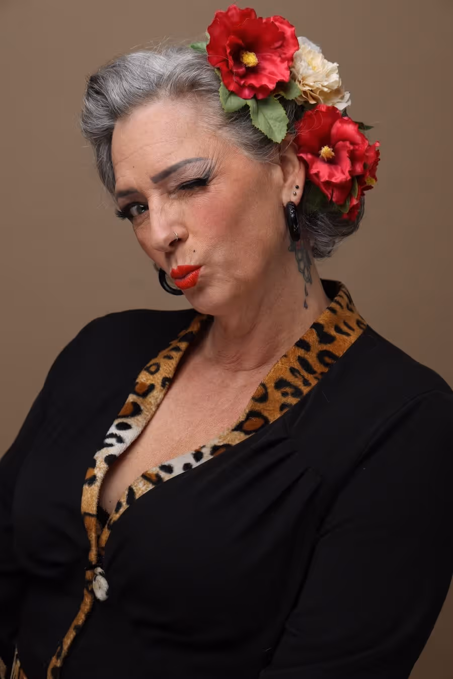 Ältere Frau mit grauen Haaren und roten Blumen im Haar zwinkert kokett, trägt schwarzen Pullover mit Leopardenmuster-Kragen.